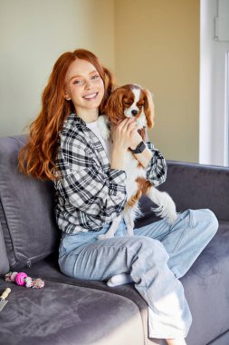 Beautiful cheerful smiling young redhead female with cute pet dog in arms at home on sofa, friendship love for pets. Pets Care. at home. indoors in living room,day time at weekends