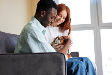 Happy black guy and caucasian lady have rest playing with dog, hugging, at home. relaxed couple enjoy weekends together, casually dressed. love, family, animals, friendship concept