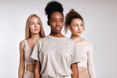 three diverse beautiful models are posing on white background. Close-up portrait of young women in casual outfit posing at camera together, looking confidently, seriously at side. copy space