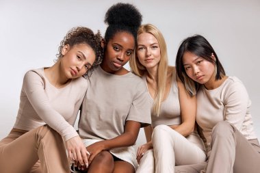 Diversity, multi-ethnic beauty concept. Four beautiful ladies of different races sit on floor posing at camera isolated in studio, African, Asian and Caucasian, with natural makeup, in row