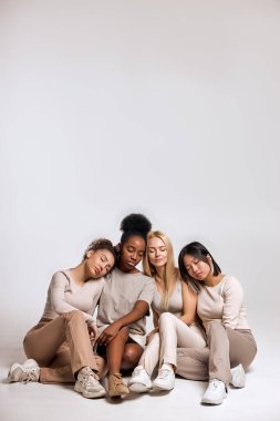 Diversity, multi-ethnic beauty concept. Group of Four beautiful women of different races sit on floor posing at camera isolated in studio, African, Asian and Caucasian, with natural makeup, in row