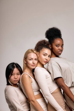 Balanced, calm diverse female models leaned on each other posing, looking at camera, isolated on beige studio background, portrait copy space. natural beauty, calmness, confidence concept