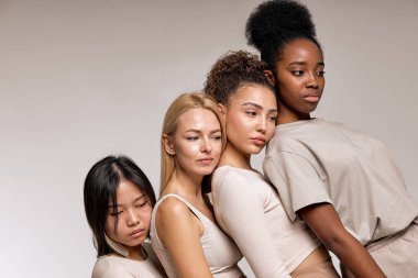 Sleepy and calm diverse women models leaned on each other posing, looking down, isolated on beige studio background, portrait copy space. natural beauty, calmness, confidence concept