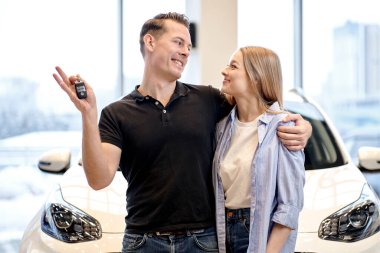 young caucasian heterosexual couple family husband and wife hugging embracing after buying new car auto at automobile dealer shop store. beautiful blonde lady and handsome guy holding key