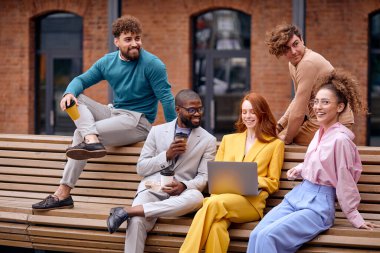 smiling multi-ethnic business colleagues employees outdoors, professionals looking positive, friendly corporate workers posing in modern downtown, sitting on bench surfing the net, drinking coffee