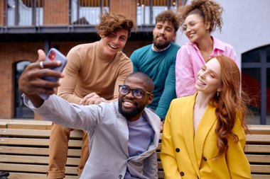 Cheerful diverse colleagues have fun making selfie on smartphone, smiling, men and women after successful cooperation meeting, dressed in fashionable stylish clothes, sitting on bench outdoors