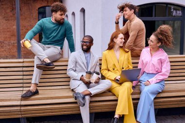 business people relaxing, talking and sitting on bench outdoors, drinking coffee, surfing the Net. caucasian and african american coworkers in trendy elegant clothes enjoy spending time outdoors