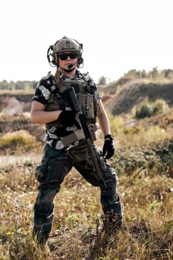 Portrait of a middle-aged bearded soldier in Woodland military uniform and cap, with headphones on head holding rifle. confident motivated military person posing at camera alone outdoors