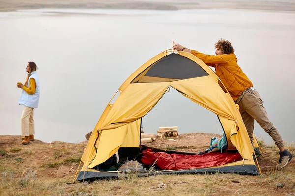couple camping in wild nature by lake river. man preparing tent and bonfire closer to the night. caucasian man and woman in casual wear in nature, woman drinking tea and enjoying the landscape