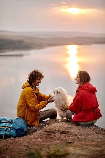 excited caucasian couple play with pet dog enjoy the lake landscape in the background, travel together, spend time together during hike, have fun, talk and smile. travel, nature, love concept