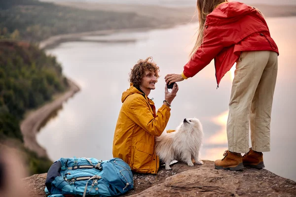 couple with pet dog Hiking in mountains together. Adventure. river in the background. Happy male and female drinking tea during hiking, travelling, spending time together outdoors in nature
