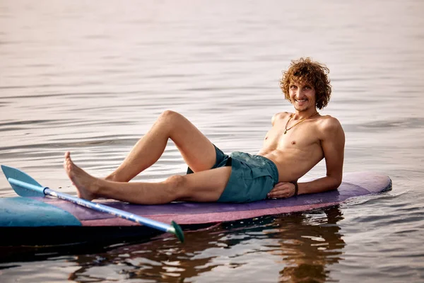 Relaxed caucasian male surfer lying on surfboard after surf on high waves. Sporty young smiling man enjoy the rest alone, looking at camera. Extreme sports, youth active lifestyle concept
