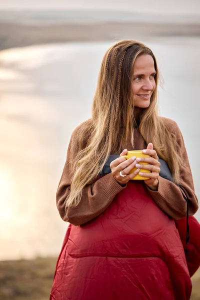 Good-looking female holding cup of tea in hands standing in contemplation of nature looking at side. attractive lady with long hair is wrapped in warm coat enjoying relax time, vacation by lake
