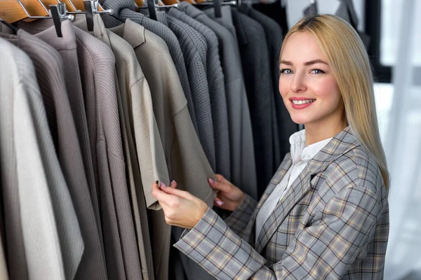 Woman looking at new collection of stylish clothes on hangers clothing rack rails, fashion design studio store concept. blonde caucasian female in jacket make choice, young customer look at camera