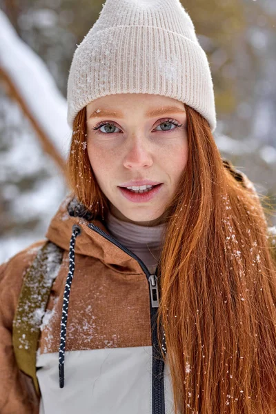 Close-up portrait of freckled redhead female posing looking at camera, on cold weather. attractive woman in beige hat having nice look, glance. snow on hair and coat. frozen cold day