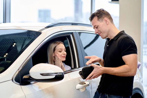 caucasian female begs boyfriend to buy new automobile in dealership, male regretfully look at empty wallet. young married couple in cars showroom, have talk, begging, woman want to get new car