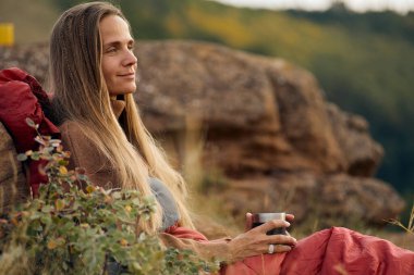 Charming woman sit leaning on big stone relaxing, drinking coffee in nature alone, enjoying landscape, dressed in casual warm sweater, looking at side in contemplation. travel, people lifestyle