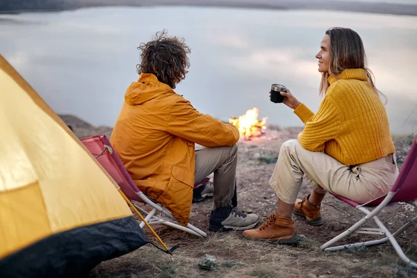 View from back on couple sitting near bonfire drinking tea, camping, travelling concept. Young caucasian man and woman in sportive clothes having conversation, spending nice time together