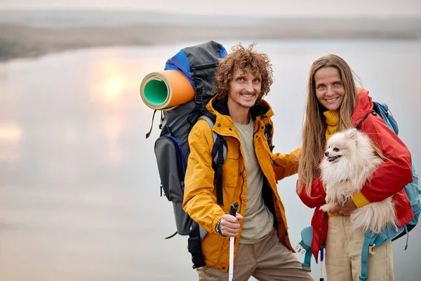 Young couple with pet dog travelling in countryside, friendly family exploring wild nature, enjoy rest together. nice caucasian man and woman with backpacks exploring new places with cute spitz dog