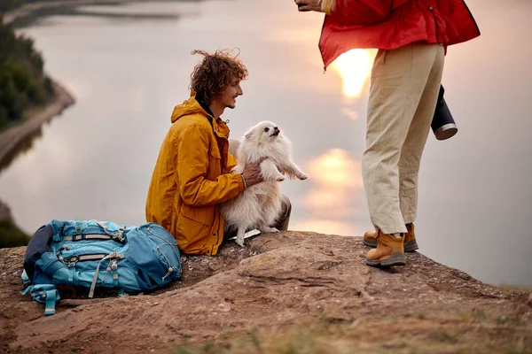 active couple with pet dog travelling in countryside, friendly family exploring wild nature, enjoy rest together. nice caucasian man and woman with backpacks exploring new places with cute spitz dog
