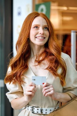 Genç bir kadın, alışveriş için aldığı kredi kartıyla mutlu. Kapalı fotoğraf, uzun kızıl saçlı, beyaz elbiseli bir kız parasını harcamayı hayal ediyor.