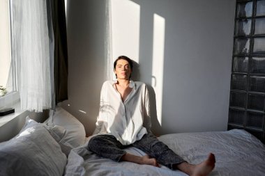Beyaz tişört giyen ve yatakta oturan yakışıklı, rahat bir adamın yakın plan fotoğrafı. Boş zaman, boş zaman, güzellik.