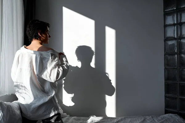 若いセクシーなモデルのバックショット彼の白いシャツを脱いで 午前中にベッドに行きます 写真の背景に横にコピースペースの影を見て裸の肩を持つペシャルな男 — ストック写真