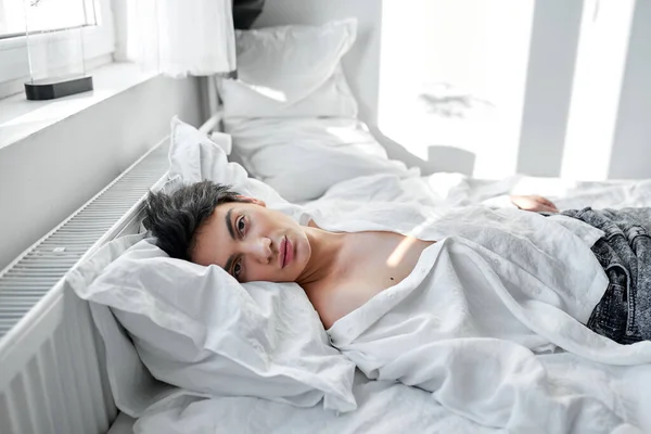 シャツを着た魅力的なブルネットの男とカメラを見ているジーンズ家で朝ベッドの上に横たわっていた 自由時間 空き時間 — ストック写真