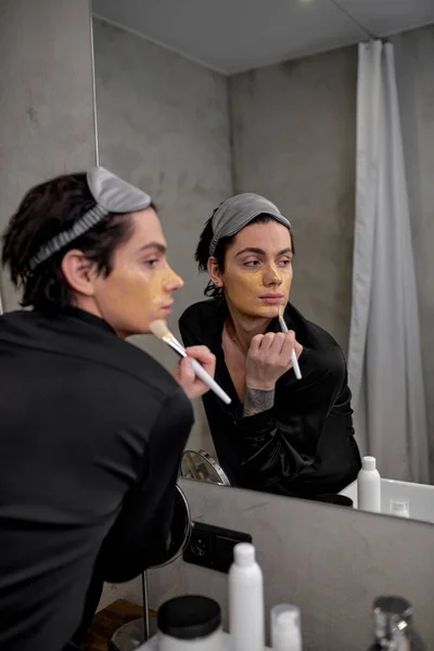 young glamour man in stylish black robe and eyemask on his head applying clay mask on face for facial treatment and skin rejuvenation in studio