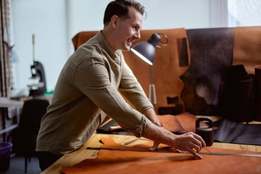 Genç, gülümseyen mutlu adam deri ürünleri elleriyle dikiyor, yan görüntüyü kapatıyor. Her türlü ticareti yapıyor.