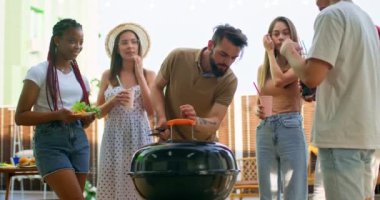 bbq Grill 'in yanında rahatlayan mutlu harika arkadaşlar. Çoklu etnik erkek kadınlar dışarıda ızgara sosis bekliyor erkekler, kızlar sohbet ediyor şişelerle konuşuyorlar kola içiyorlar yavaş çekim kutlamaları yapıyorlar.