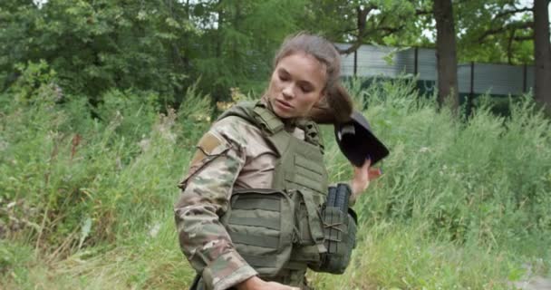 緑の森のスローモーションに立ってキャップ ボディアーマー ヘッドセットを離陸されている自信を持って若いアメリカの兵士の女性のクローズアップビュー — ストック動画