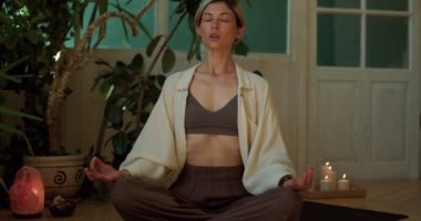 Nilüfer çiçekli güzel, açık renk saçlı bir kadın evde kapalı gözlerle oturmuş yerde tek başına meditasyon yapıyor. Kadın derin nefes alma egzersizleri yapıyor..