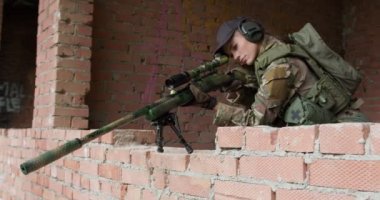 Kamuflajlı ciddi bir askeri kadın, keskin nişancı tüfeğiyle terk edilmiş bir binanın penceresinden nişan alıyor. Saldırı tüfekli bir kadın, silahlı bir kız çocuğu.