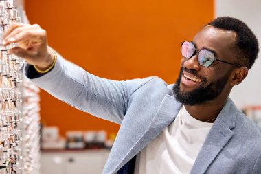 Neşeli bir adam gözlük almayı eğlenceli bir uğraş olarak görüyor. Eğlence, yakın plan fotoğraf. Afro mağaza asistanı, alıcı müşteriler için yeni moda gözlük koleksiyonu sunuyor.