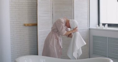 Genç, gülümseyen, neşeli bir kadın kafasına ıslak saçlarıyla beyaz bir havlu koyup banyoda ya da otelde küvetin arkasında duruyor. Yavaş hareket. Kız kolunu suya sokuyor, oturuyor.