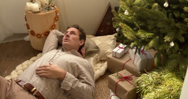 カジュアルなシャツの床の残りの部分を楽しんで横にあるリラックスした白人男性は クリスマスツリーの横にある 良い気分の写真リラックスした男性は自由な時間を楽しんで新年を祝う家で陽気なクリスマス — ストック動画