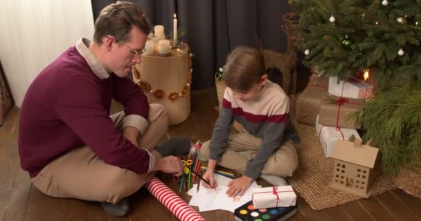 快乐的千禧爸爸和可爱的小儿子一起画画 坐在装饰过的圣诞树旁 在家里共度佳节 共度佳节 — 图库视频影像