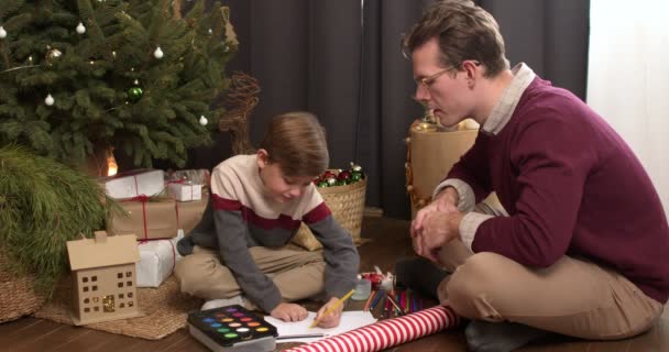 友善的爸爸和儿子在准备圣诞节的庆祝活动 画在圣诞树附近的地板上 在圣诞快乐的日子里 愉快而快乐 照片为2022年及圣诞快乐概念 — 图库视频影像