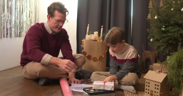 小男孩为心爱的人准备圣诞惊喜 坐在地板上画画 和蔼可亲的高加索人帮助和儿子聊天 在温暖的地板上 一起度假 庆祝新年 — 图库视频影像