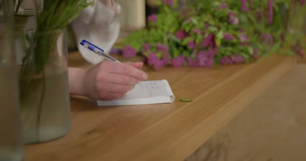 仕事でノートを作る花屋の女性のクローズアップ手ノートやテーブルの上に横たわる花で書く ビジネス 販売および花粉の概念 ペンを持ち注文をすることに集中して — ストック動画