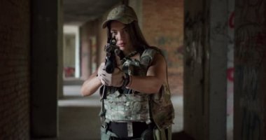 Silahlı genç kız, silahlı çatışma bölgesinde kamuflaj üniforması içinde kapalı alanda ağır çekimde saldırı tüfeği hedefliyor. Rus-Ukrayna savaşı. Silahla nişan alıyor, atıcı hedef paintball 'u arıyor.