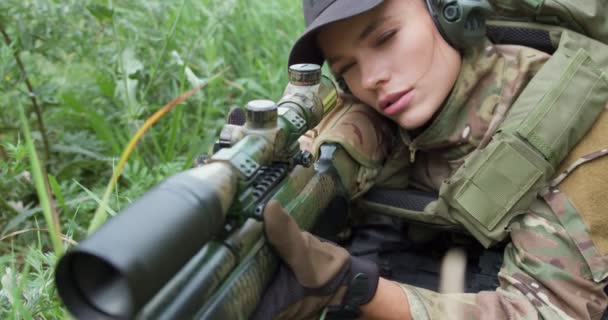 头戴帽子 头戴耳机 带着狙击步枪的战术装备的严肃女人坐在草地上 训练用枪射击 慢动作侧视 — 图库视频影像