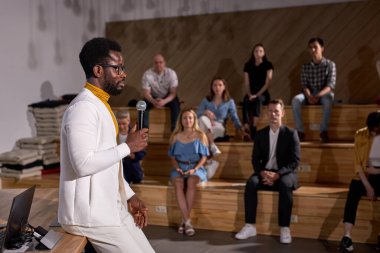 Kafkas grubuna mikrofonla ders veren Afro öğretim görevlisi. Genç siyahi öğretmen konferans salonunda finans hakkında konuşuyor. Gözlüklü ve beyaz giysili yakışıklıya yan bakış