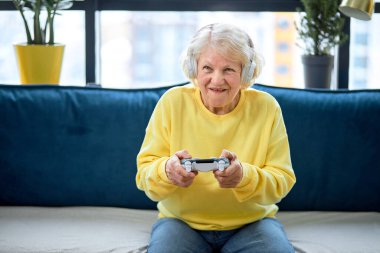 Sanal savaştan zevk alan, oyuna odaklanmış yaşlı bir kadın. Ev eğlencesi kavramı. Gündelik giyimli komik beyaz kadın evde kanepede oturmuş video oyunu oynuyor.