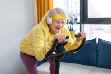 70 'li yaşların ortasında yaşlı bir kadın evde staj bisikleti üzerinde egzersiz yapıyor. Aktif, yetişkin bir kadın spor kıyafetleri içinde oturma odasında bisiklet egzersizi yapıyor. aktif emeklilik yaşam tarzı kavramı