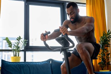 Egzersiz bisikleti ya da evde tek başına bisiklet süren kendine güvenen siyah bir adam. Çıplak gövdeli fitness adamı kardiyo eğitimi alıyor. Spor, egzersiz, sağlıklı yaşam tarzı konsepti.