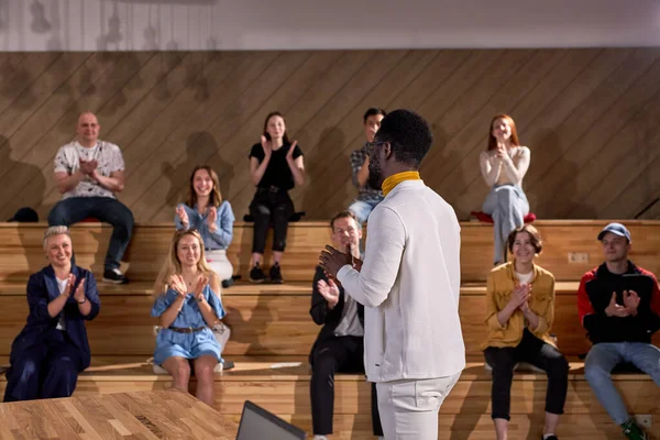 Erkek Afro-Amerikan konuşmacı konferans salonunda sunum yapıyor, izleyiciler. Arka plandaki genç katılımcılar el çırpıyor. Konferans etkinliği, eğitim. Eğitim, çeşitlilik, kapsayıcı kavram.