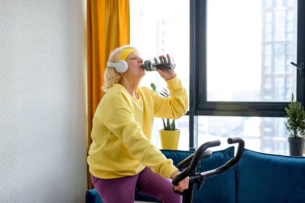 Elderly person training on stationary bicycle doing physical exercise and activity. Senior caucasian woman using cardio cycling machine to train legs muscles with gymnastics at home.