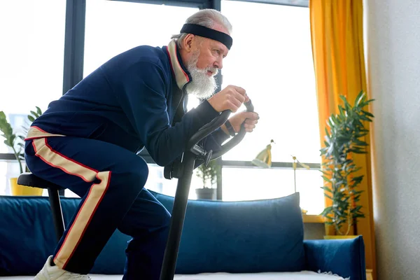 Elderly person training on stationary bicycle doing physical exercise and activity. Senior caucasian man using cardio cycling machine to train legs muscles with gymnastics at home.